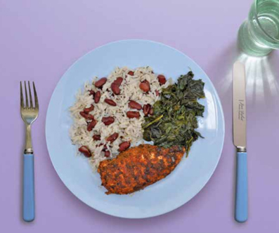 Jerk chicken on a plate with rice and peas and callaloo. There is a glass of water and cutlery on the table. 