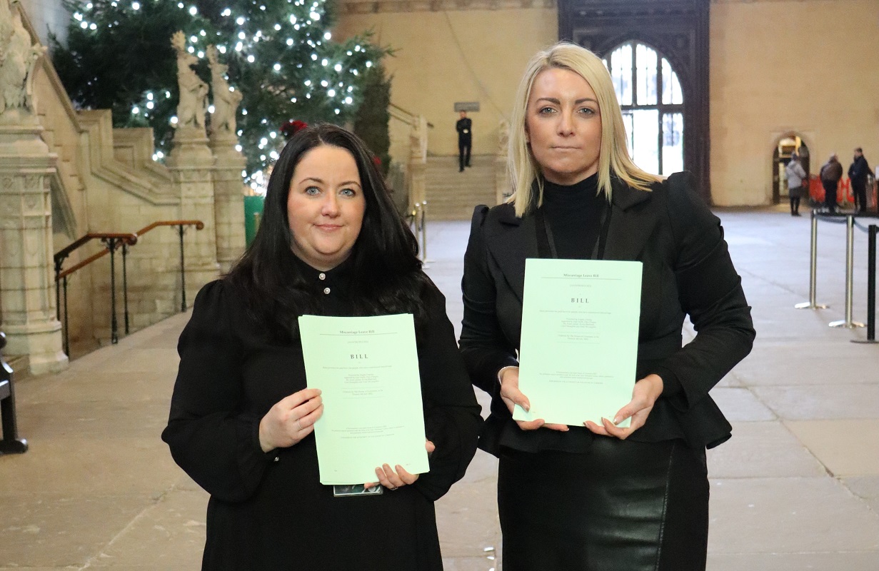 Angela Crawley and Keeley wearing black stand in the lobby of the House of Commons holding paper copies of the Miscarriage Leave Bill