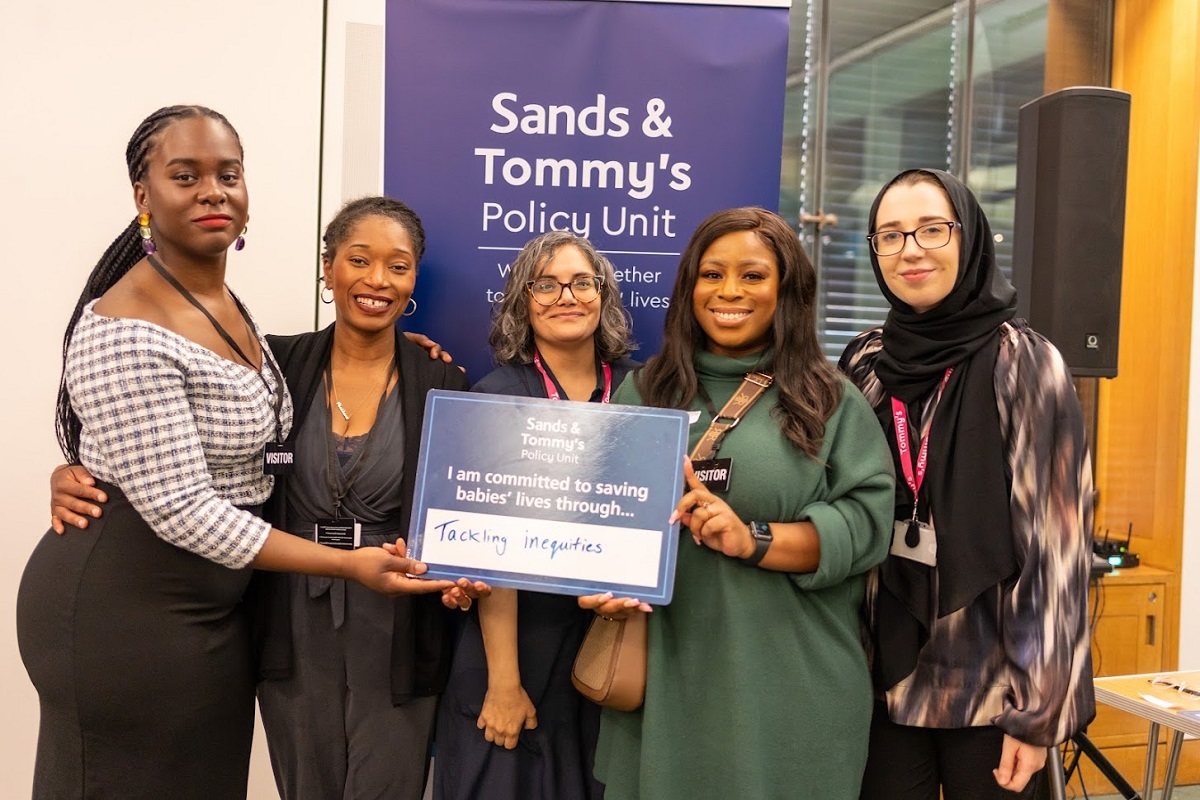 A group of Tommy's staff and ambassadors standing in front of a JPU banner holding a sign which says "I am committed to saving babies' lives through tackling inequities"