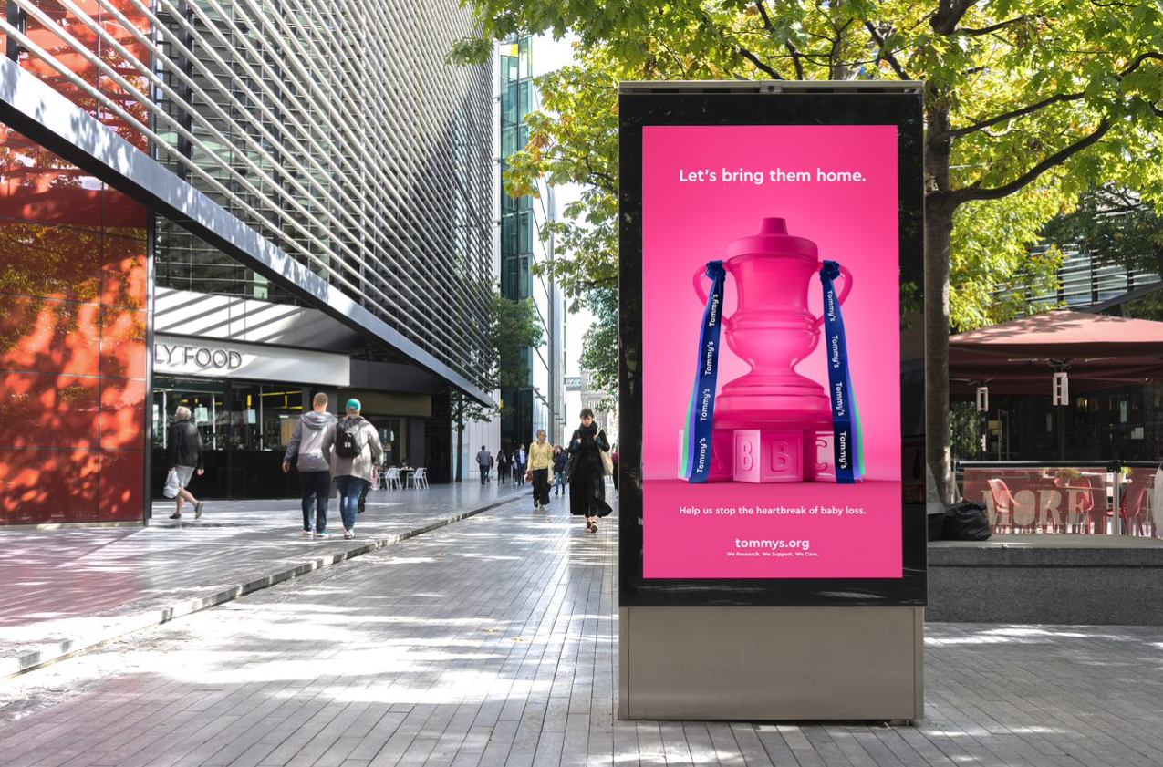 A pink Tommy's ad on a bus shelter digital screen showing baby tippy cup styled like the FA Cup trophy